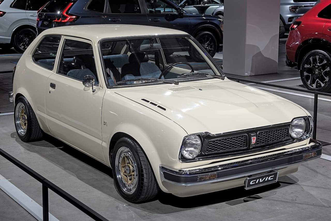 White 1972 Honda Civic hatchback on display platform in modern car showroom surrounded by contemporary vehicles and concrete columns