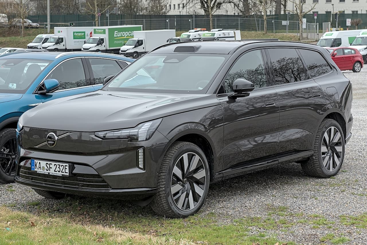 Dark gray Volvo EX90 electric SUV with German license plate parked on gravel with Europcar vans in background