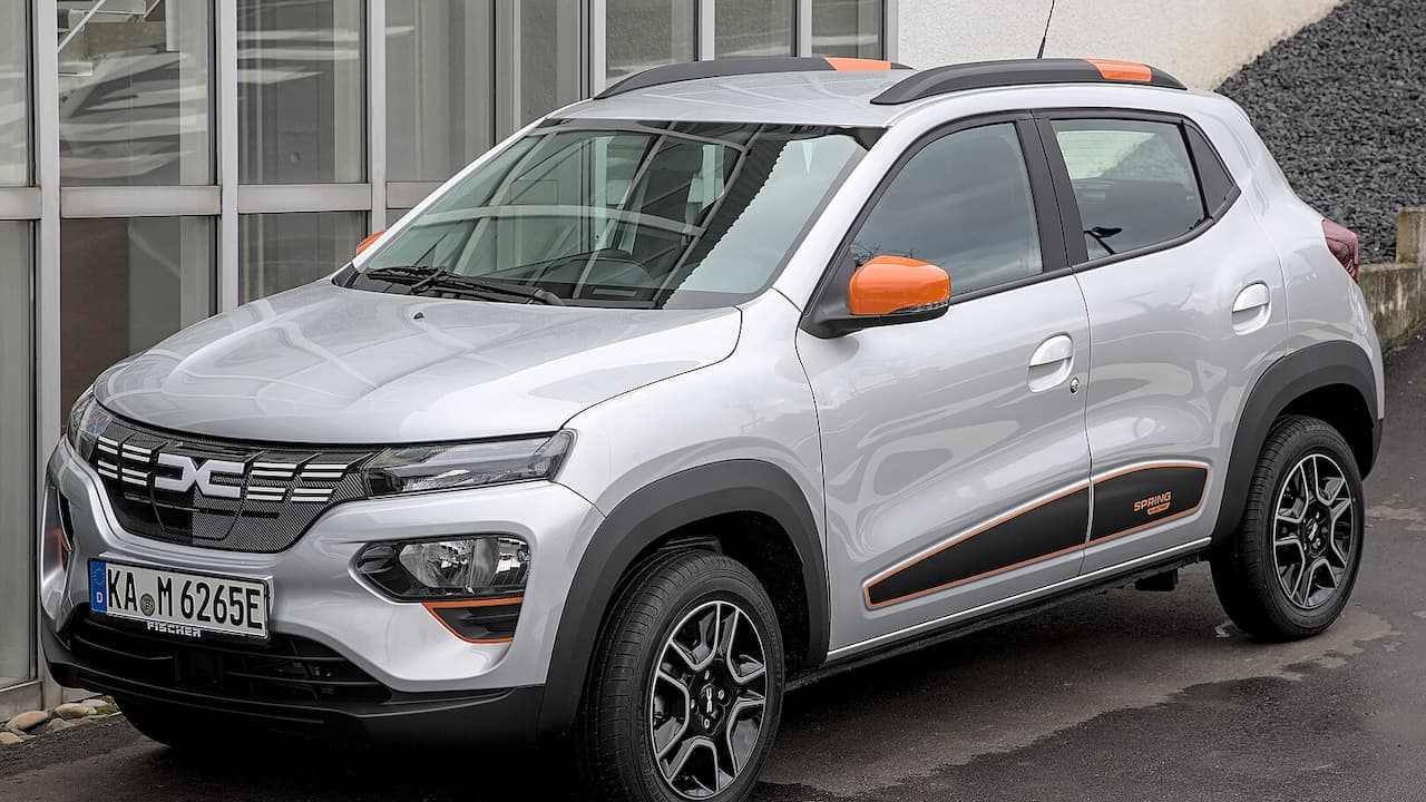 Silver Dacia Spring with orange accents parked outside modern building, featuring distinctive Dacia grille and compact SUV styling