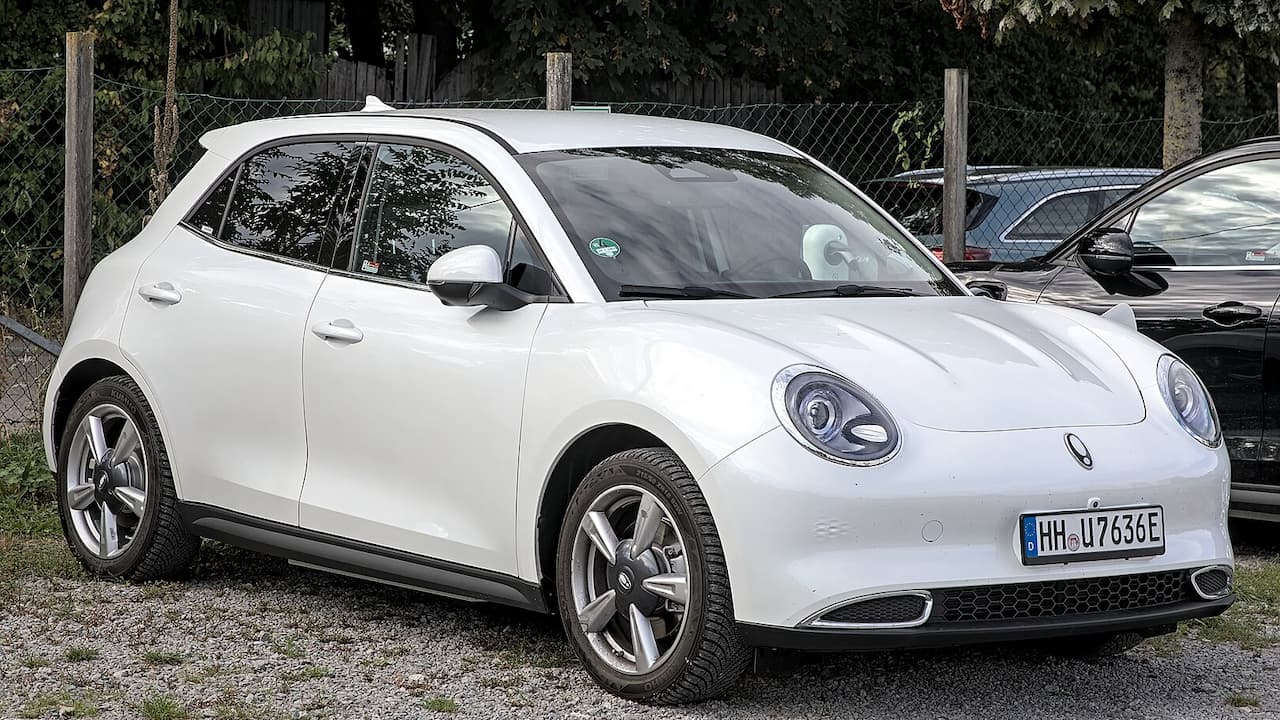 White Ora Funky Cat electric vehicle with round headlights and German license plate parked near chain-link fence