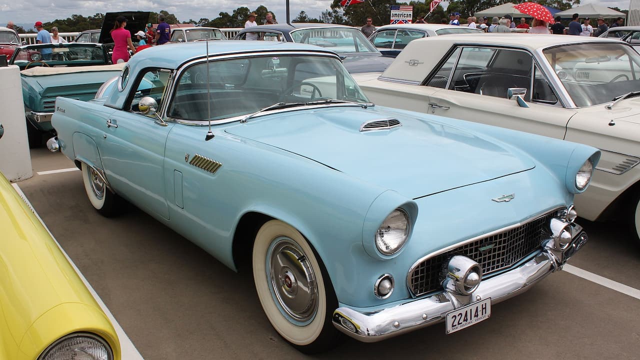 Light blue 1956 Ford Thunderbird with white hardtop and whitewall tires at classic car show with crowds