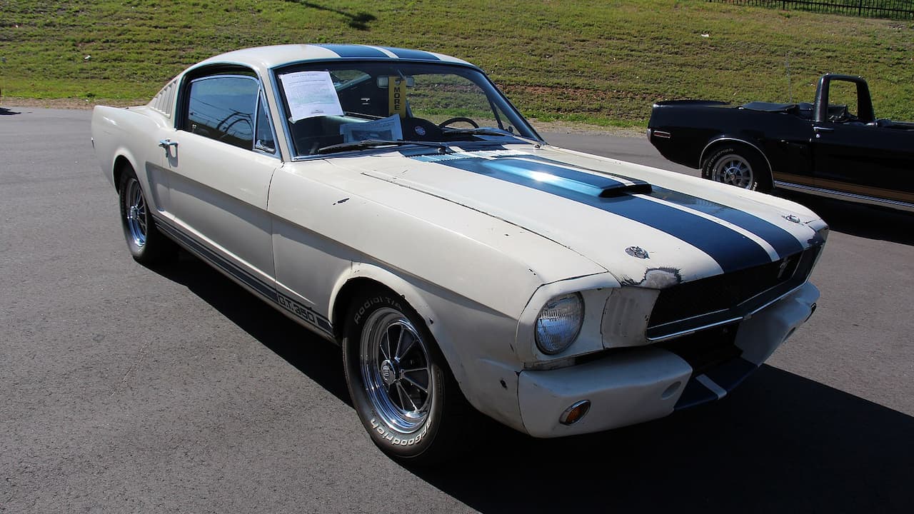 White 1965 Shelby Mustang GT350 with blue racing stripes and chrome wheels parked on asphalt at car show