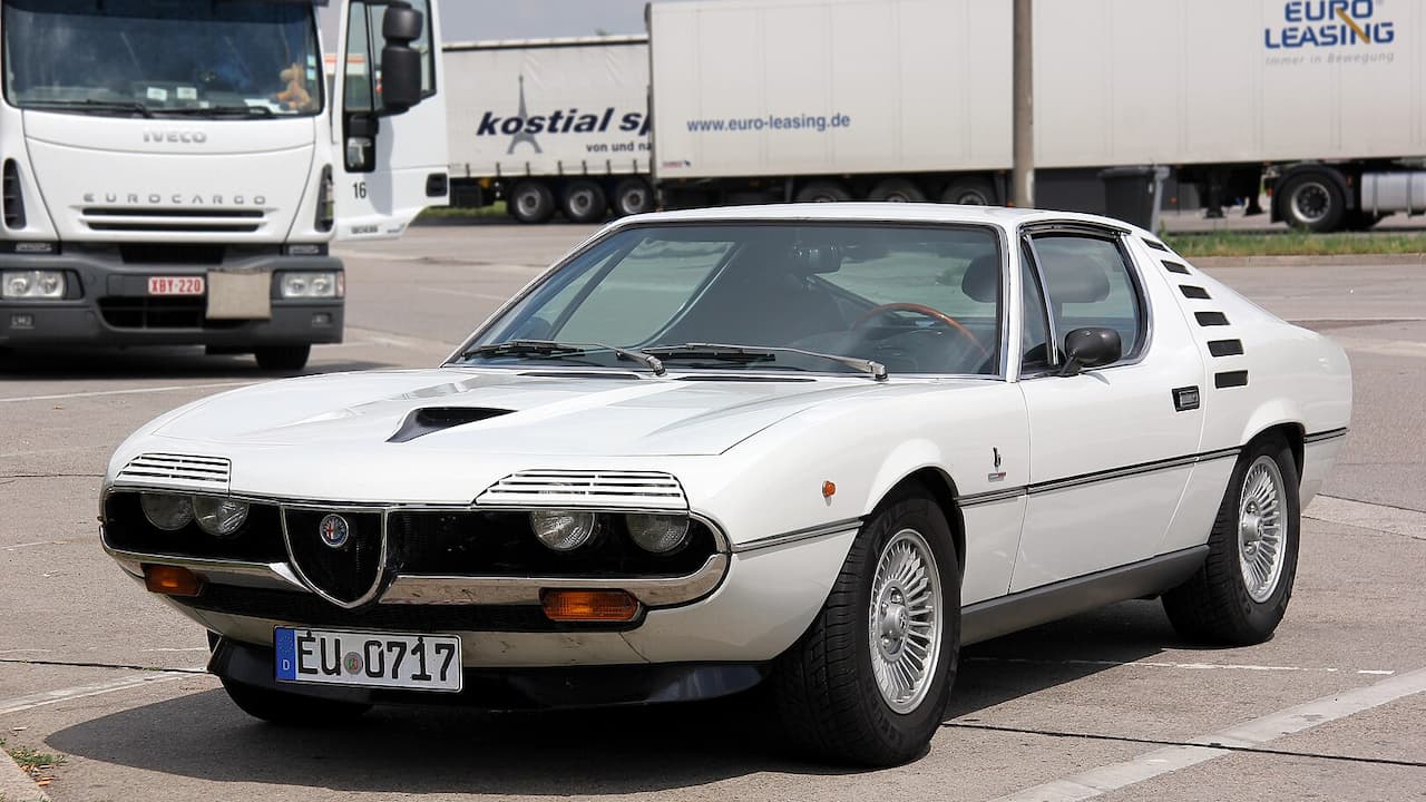 White 1970 Alfa Romeo Montreal sports car with distinctive front grille parked near commercial trucks at industrial location
