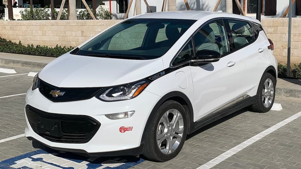 White Chevy Bolt EV electric hatchback parked in paved lot with modern building and landscaping in background