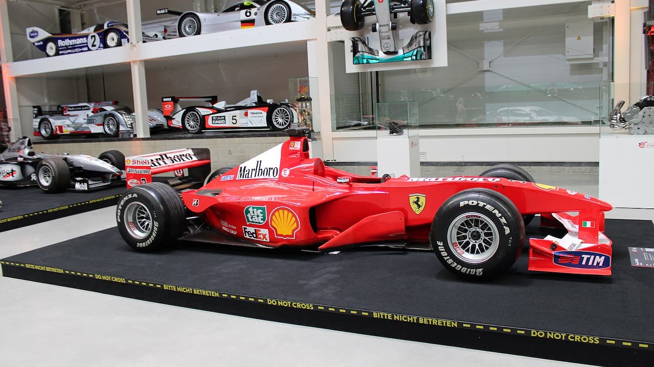 Red Ferrari F1-2000 Formula One race car displayed in museum with other racing cars on shelves behind