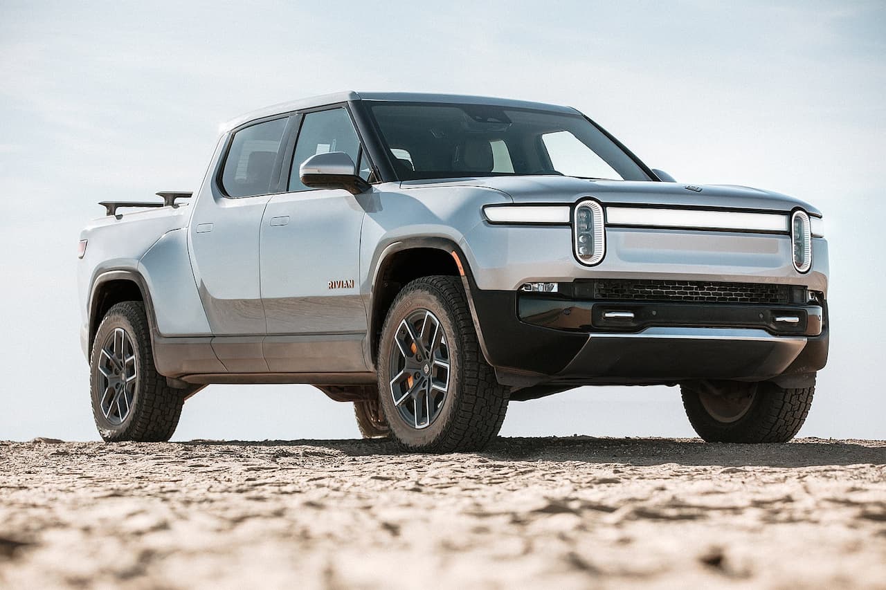 Silver Rivian R1T electric pickup truck parked on sandy terrain with distinctive LED light bar and rugged all-terrain tires