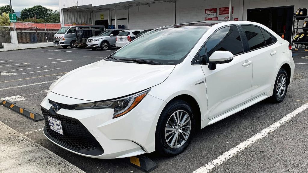 White Toyota Corolla Hybrid parked in parking lot with other vehicles and service station building visible