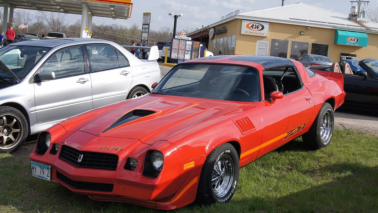 Red 1979 Chevrolet Camaro Z/28 with black racing stripes parked at gas station among other vehicles
