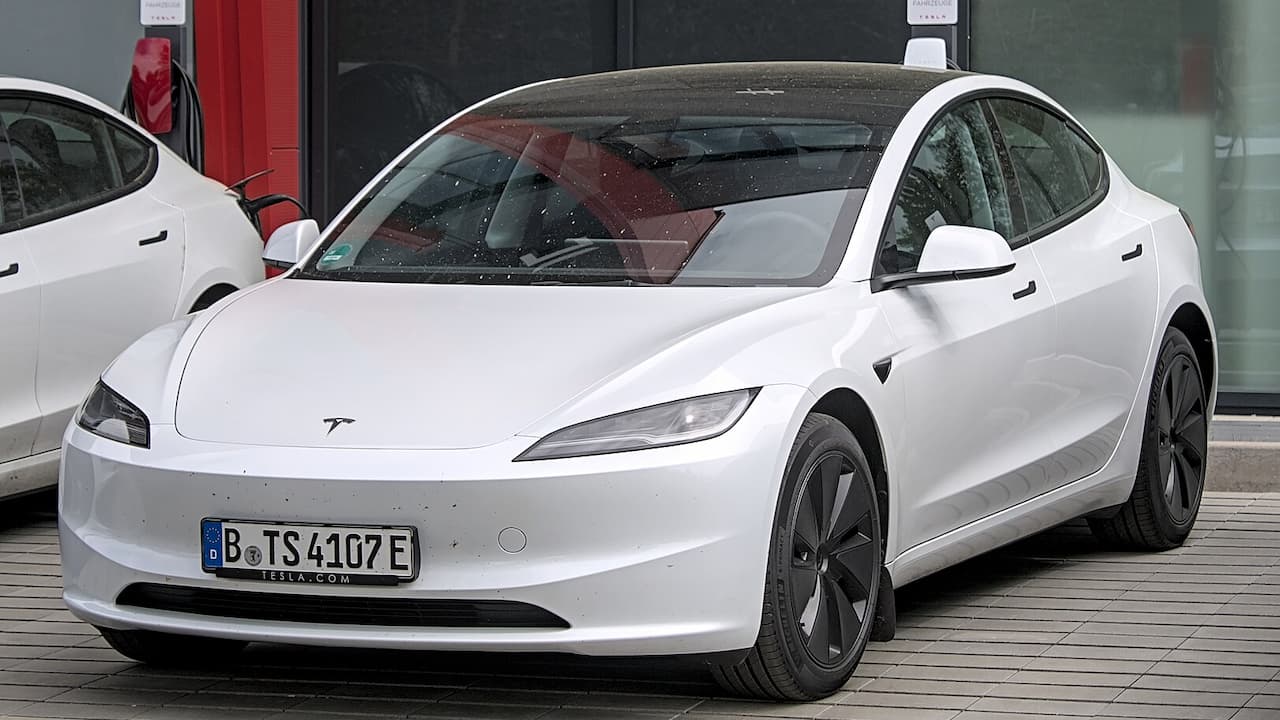 White Tesla Model 3 parked on brick pavement with distinctive minimalist design, blue license plate, and building background