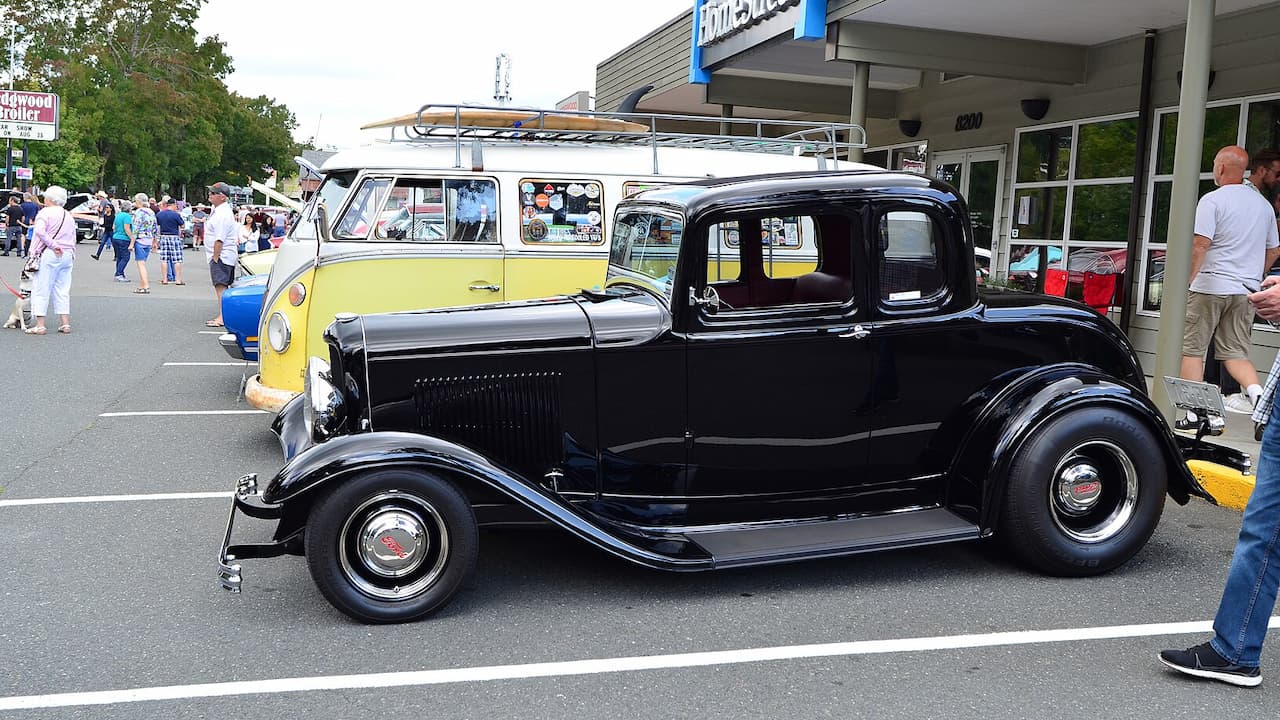Black 1932 Ford 5-Window Coupe displayed at classic car show with yellow Volkswagen van and spectators in background