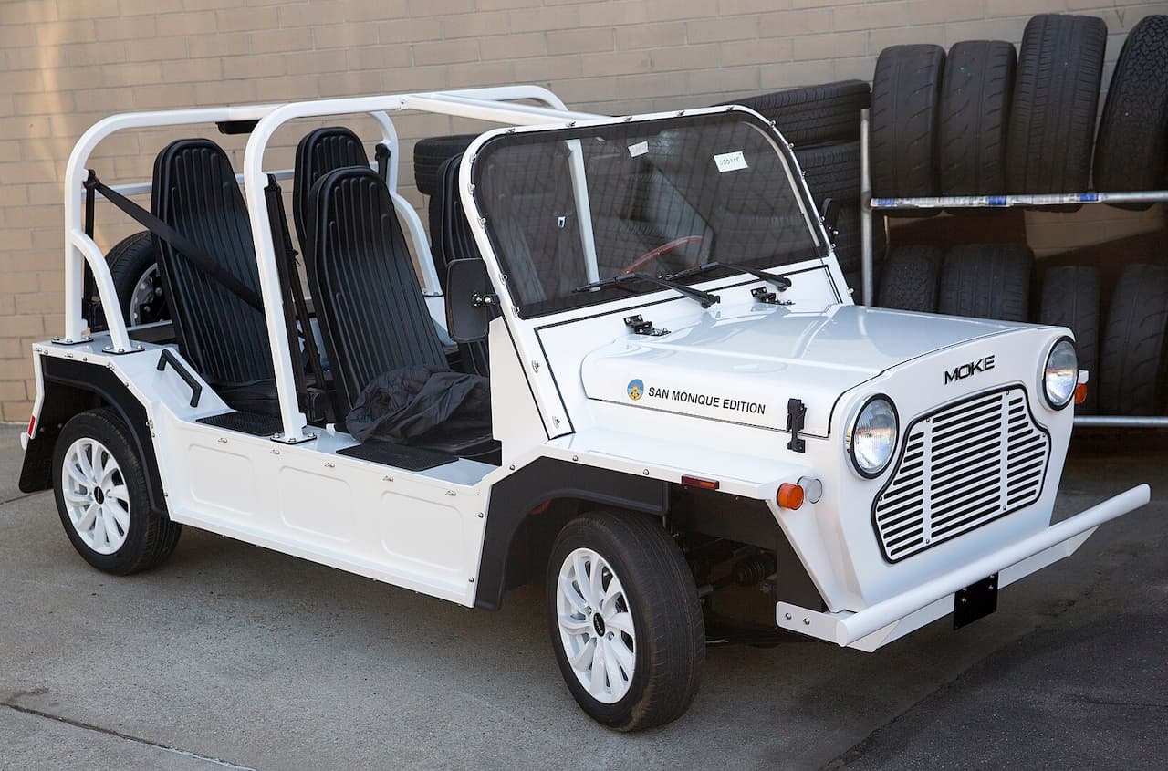 White Moke America electric vehicle with open sides, black striped seats, roll cage, parked outside building with tires stacked