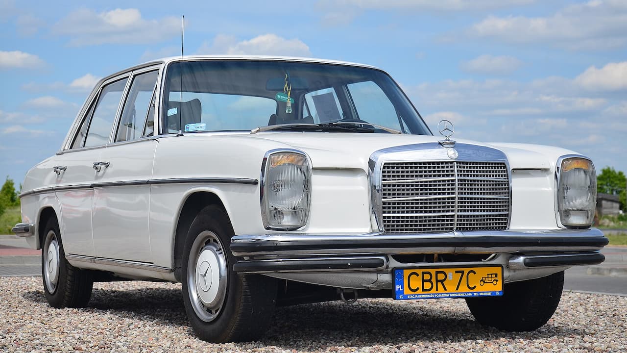 White vintage Mercedes-Benz W115 diesel sedan with classic grille, round headlights, and European license plate parked on gravel surface