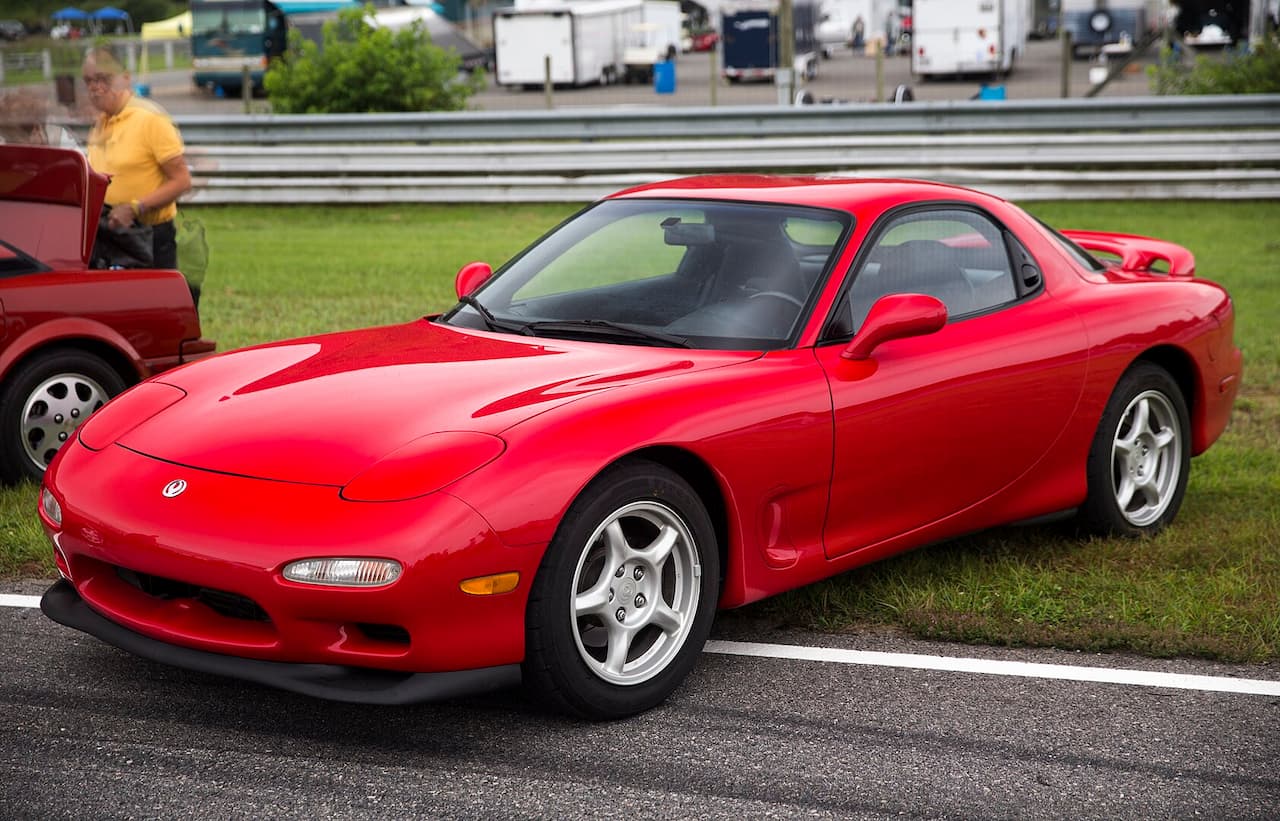 Red Mazda RX-7 sports car with pop-up headlights parked on asphalt at outdoor event with spectators and vehicles
