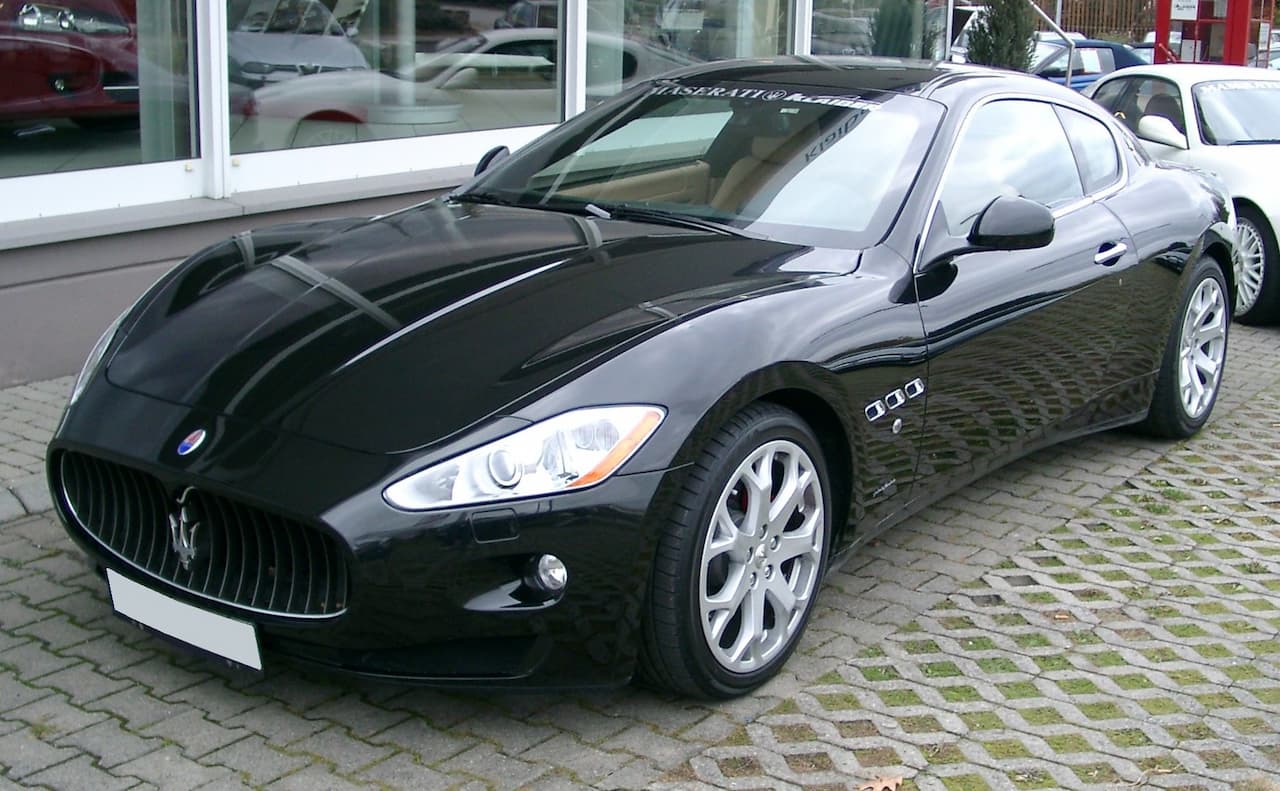 Black Maserati GranTurismo sports car parked on cobblestone street in front of car dealership with glass windows