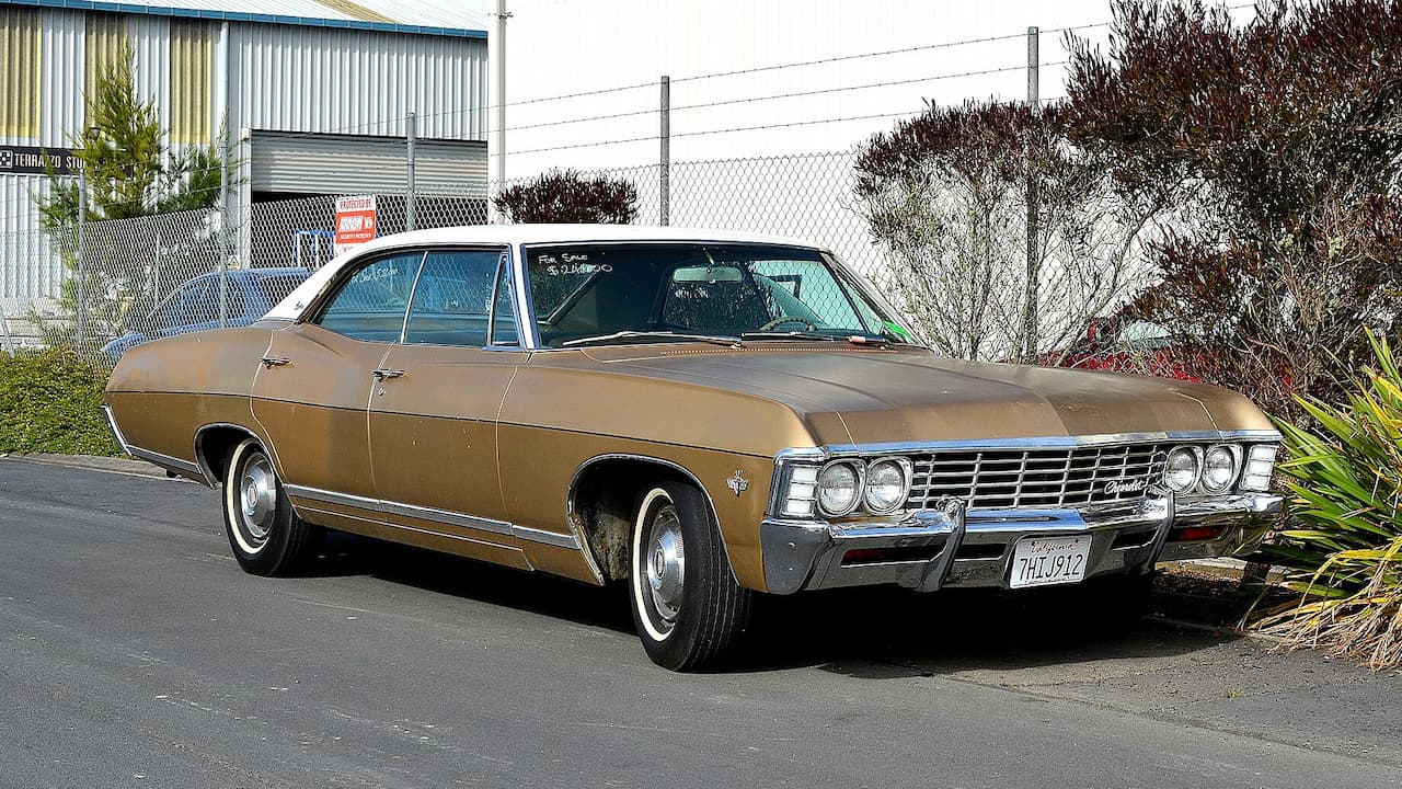 Bronze 1960s Chevrolet Caprice classic muscle car with white roof parked on asphalt near chain-link fence and industrial buildings