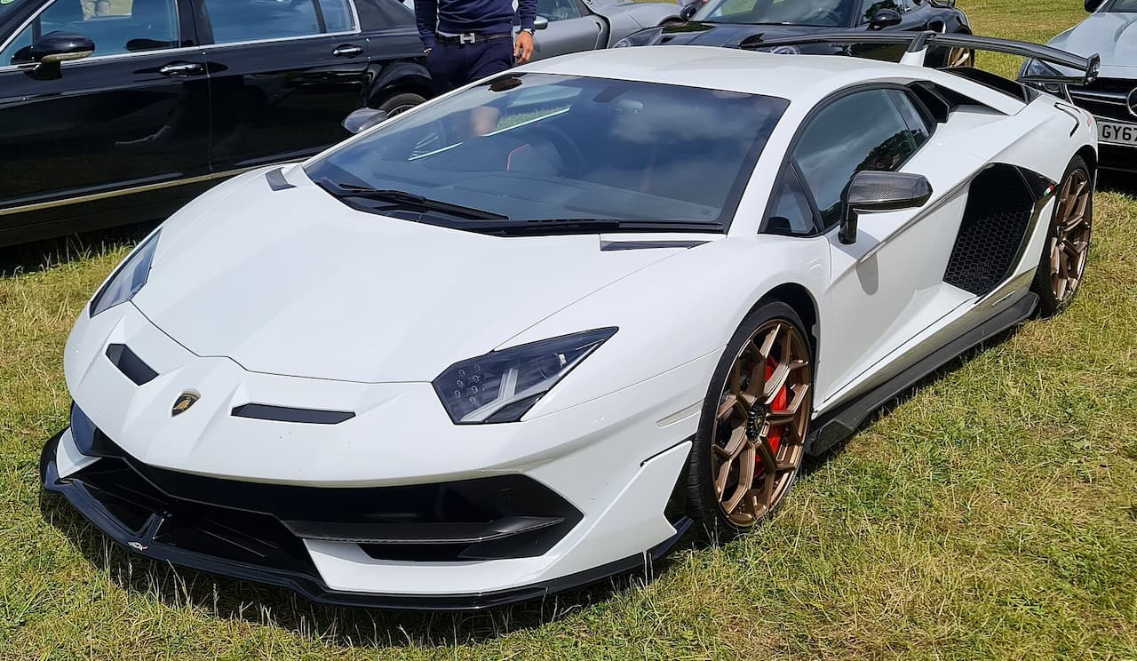 White Lamborghini Aventador SVJ supercar with active suspension systems, angular design and bronze wheels parked on grass at car show event