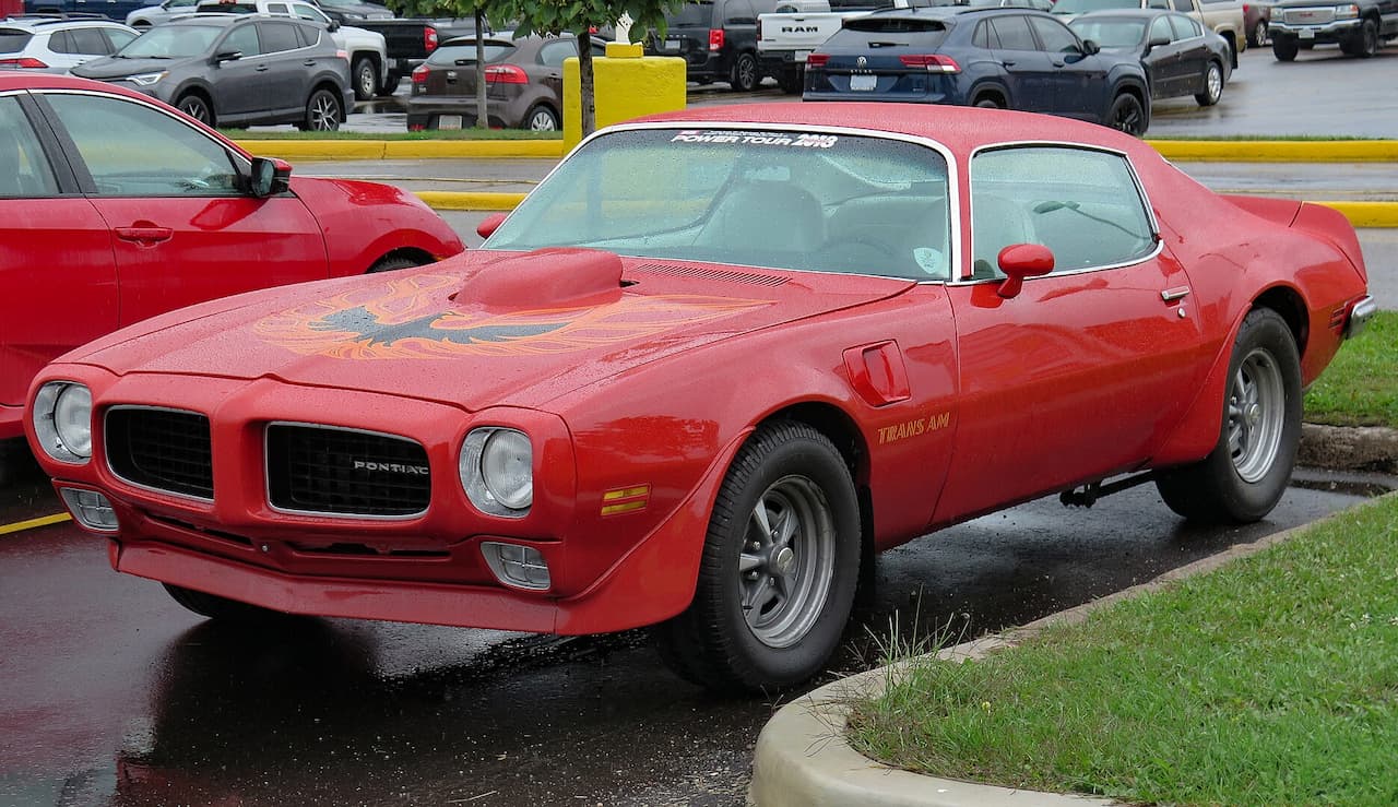 Red 1973 Pontiac Firebird Trans Am muscle car with hood bird decal parked on wet pavement at car show