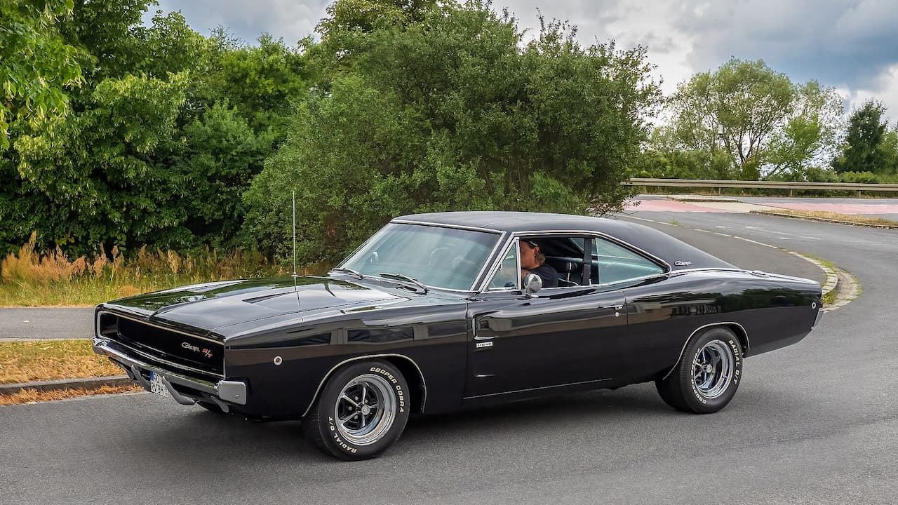 Black 1970 Dodge Charger R/T with chrome wheels and hood scoops driving on rural road with trees