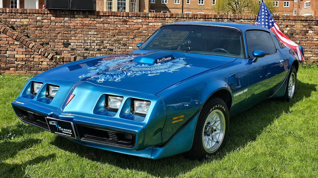 Blue Pontiac Firebird Trans Am with eagle hood graphics and American flag displayed on grass near brick wall