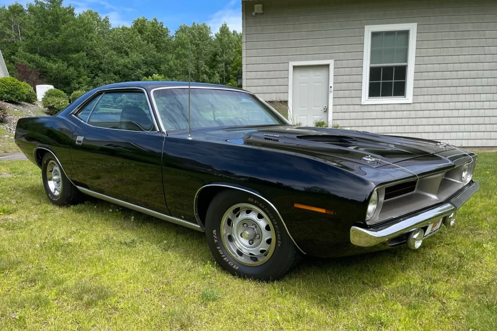 Black 1970 Plymouth Barracuda with quarter-panel scoops parked on grass near white house with trees