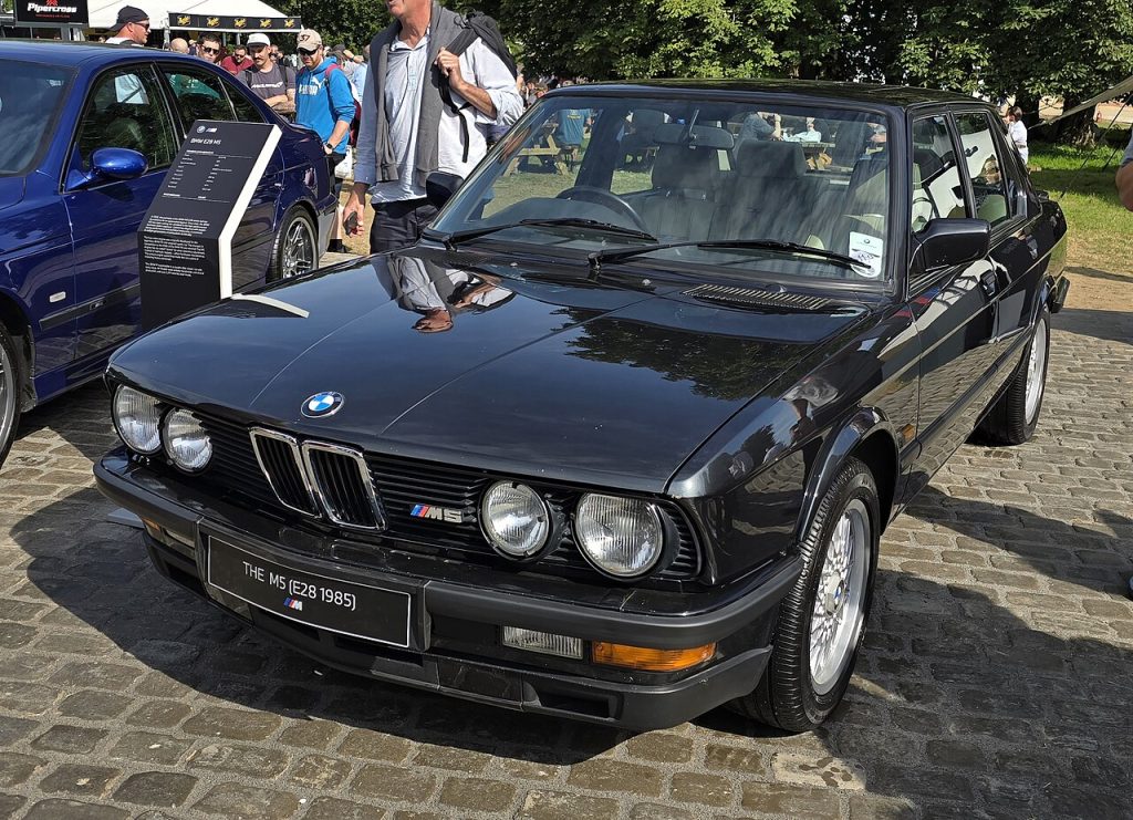 Black 1985 BMW M5 parked at outdoor car show with people gathered around other classic vehicles in background