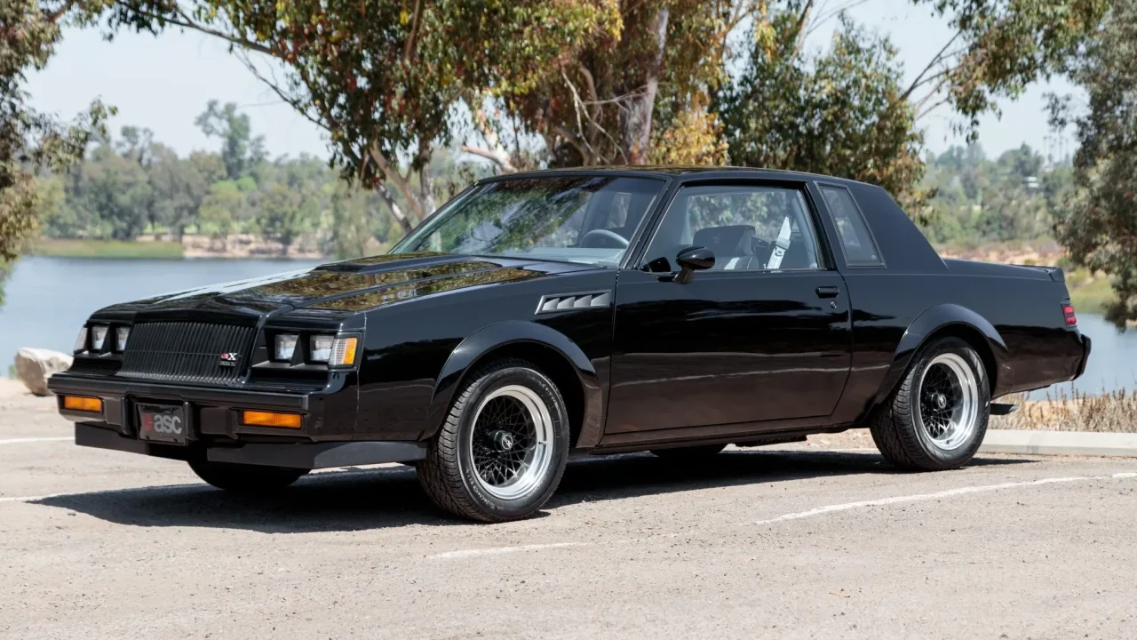 Black 1987 Buick GNX muscle car with distinctive wheels parked on pavement near trees and water