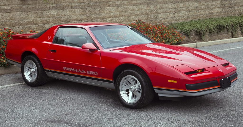 Red 1988 Pontiac Firebird V8 parked on asphalt road with stone retaining wall and grassy hill in background
