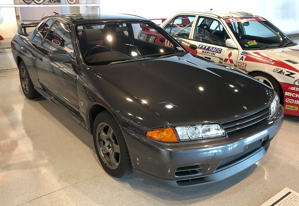 Dark gray 1989 Nissan Skyline R32 GT-R displayed indoors at car show with other vehicles visible in background