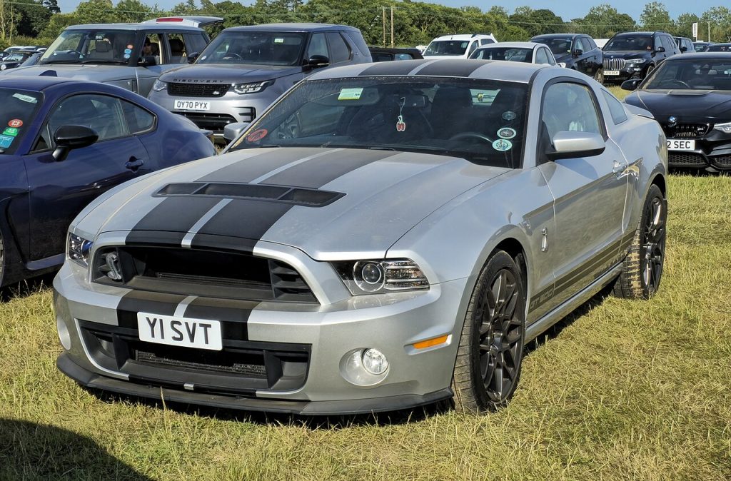 Silver 2013 Ford Mustang Shelby GT500 with black racing stripes parked on grass at car show event