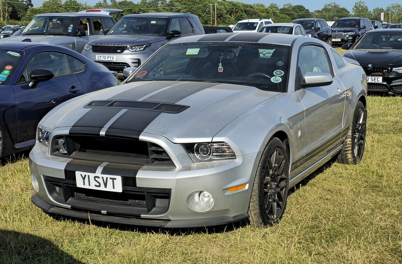 Silver 2013 Ford Mustang Shelby GT500 with black racing stripes parked on grass at car show event