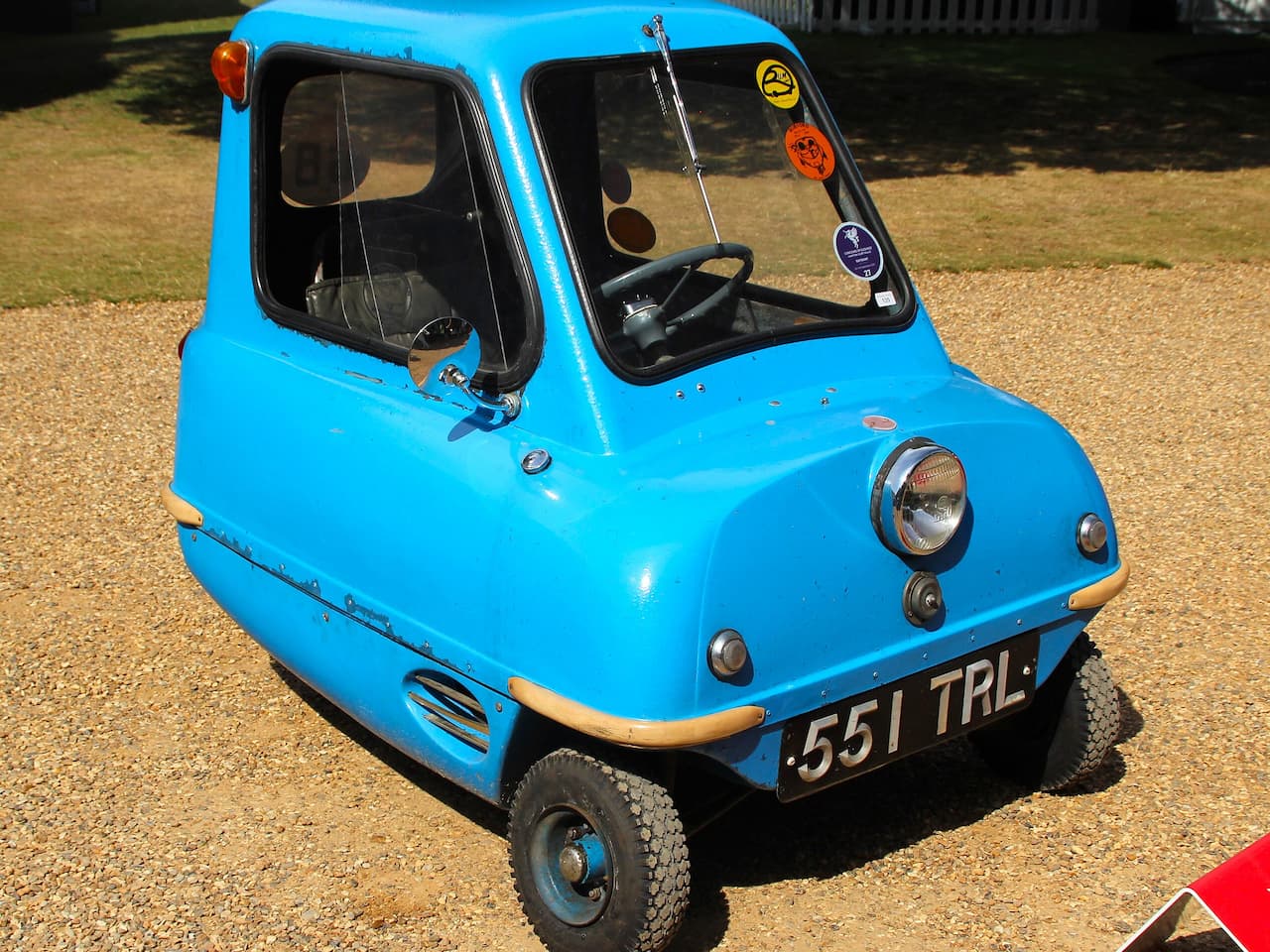 Blue Peel P50 microcar with license plate "551 TRL" parked on gravel surface in sunny outdoor setting