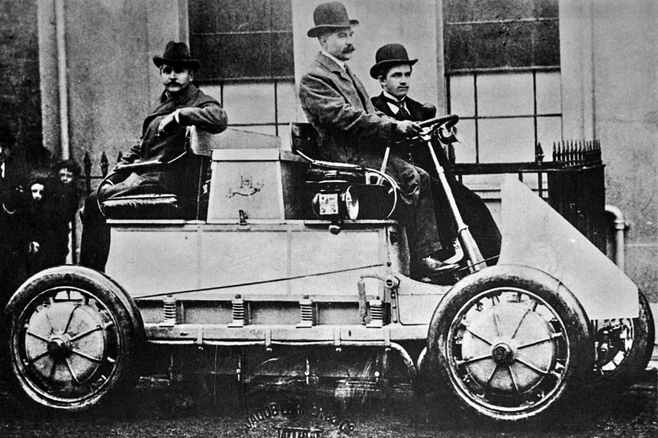 Black and white 1900 photograph of three men in bowler hats aboard Ferdinand Porsche's hybrid Lohner-Porsche electric vehicle