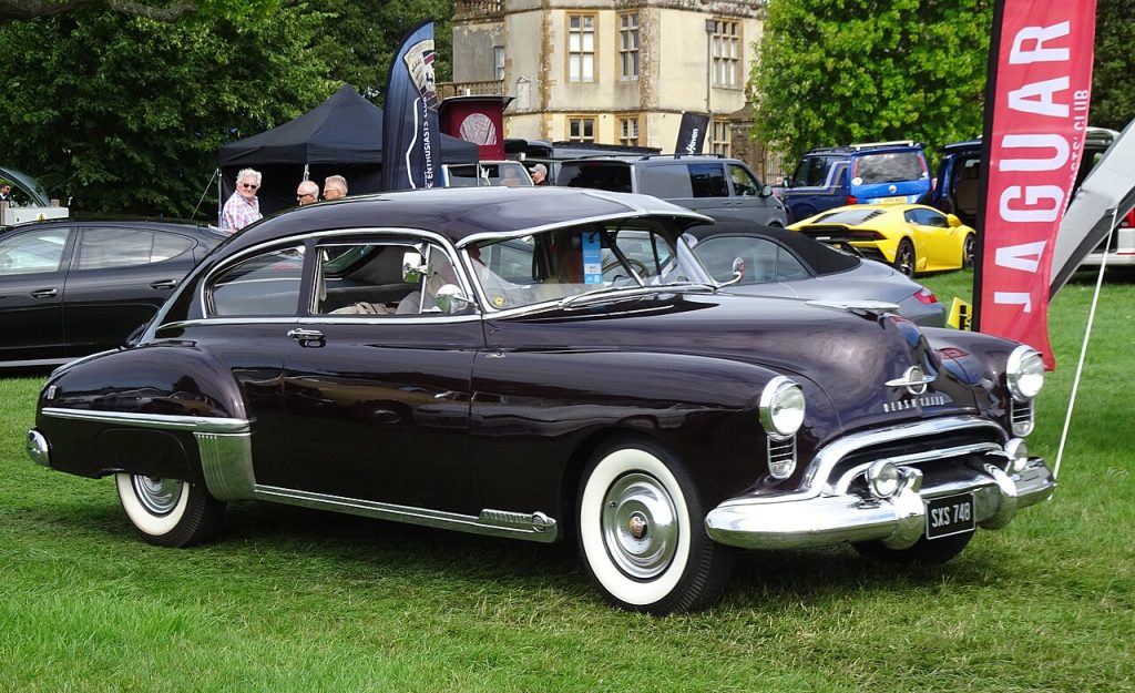 Black 1949 Oldsmobile Rocket 88 classic car displayed on grass at outdoor car show with people nearby