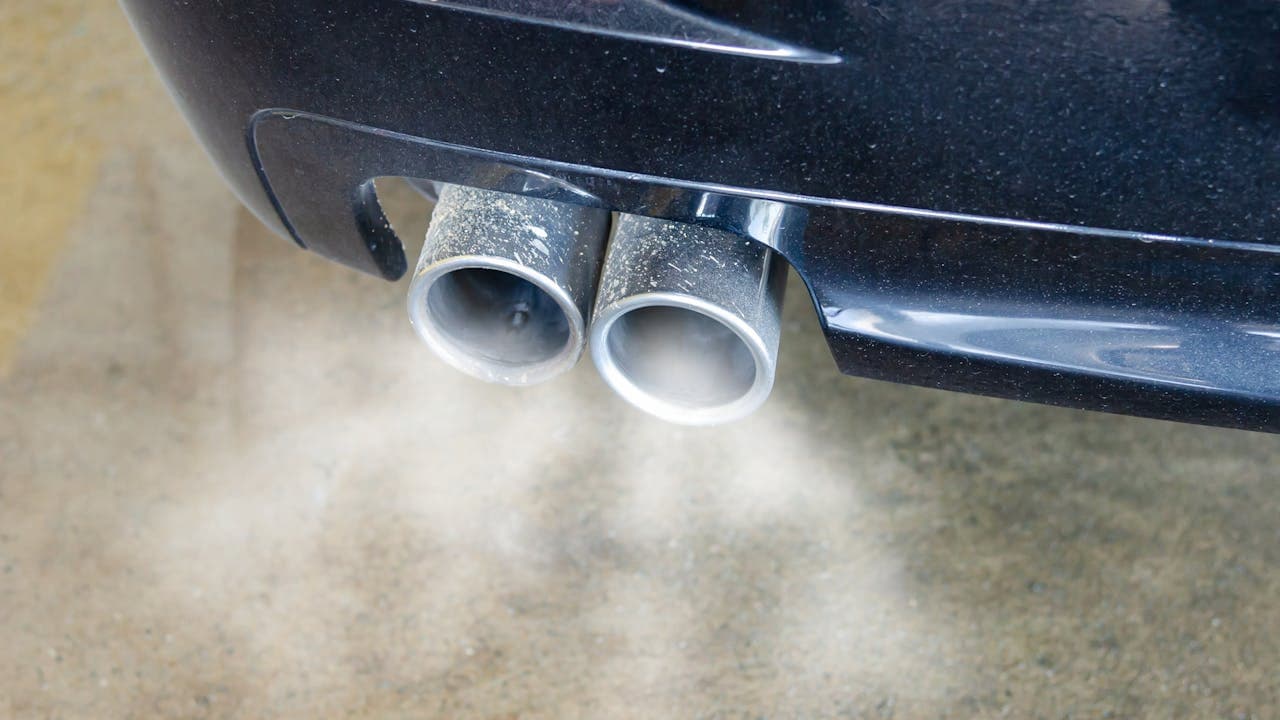 Car exhaust pipes emitting white smoke onto concrete pavement, showing vehicle emissions from dual tailpipes