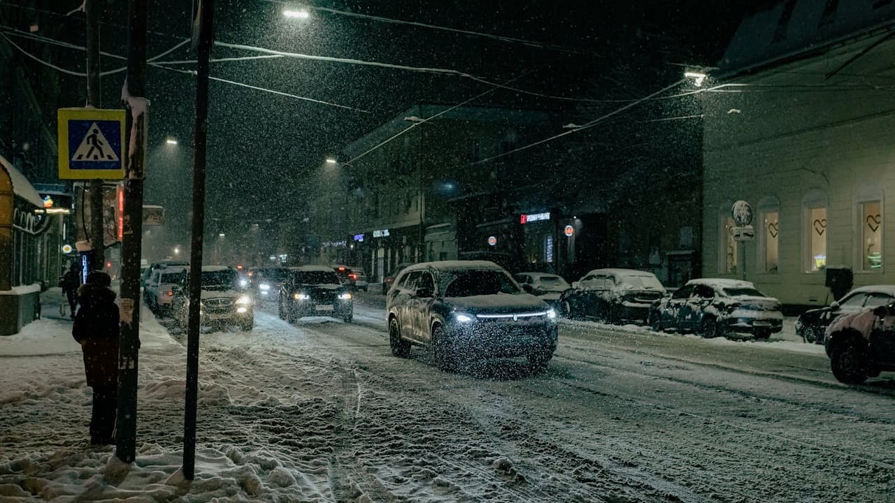 Snowy city street at night with cars driving through heavy snowfall, street lights illuminating the winter scene with pedestrians waiting