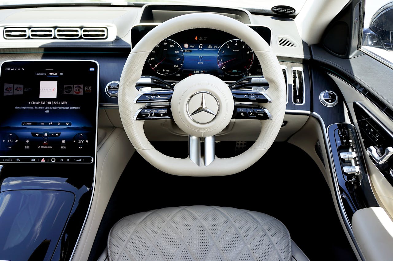 Mercedes-Benz S-Class interior showing white steering wheel, digital dashboard, large touchscreen, and quilted white leather seats