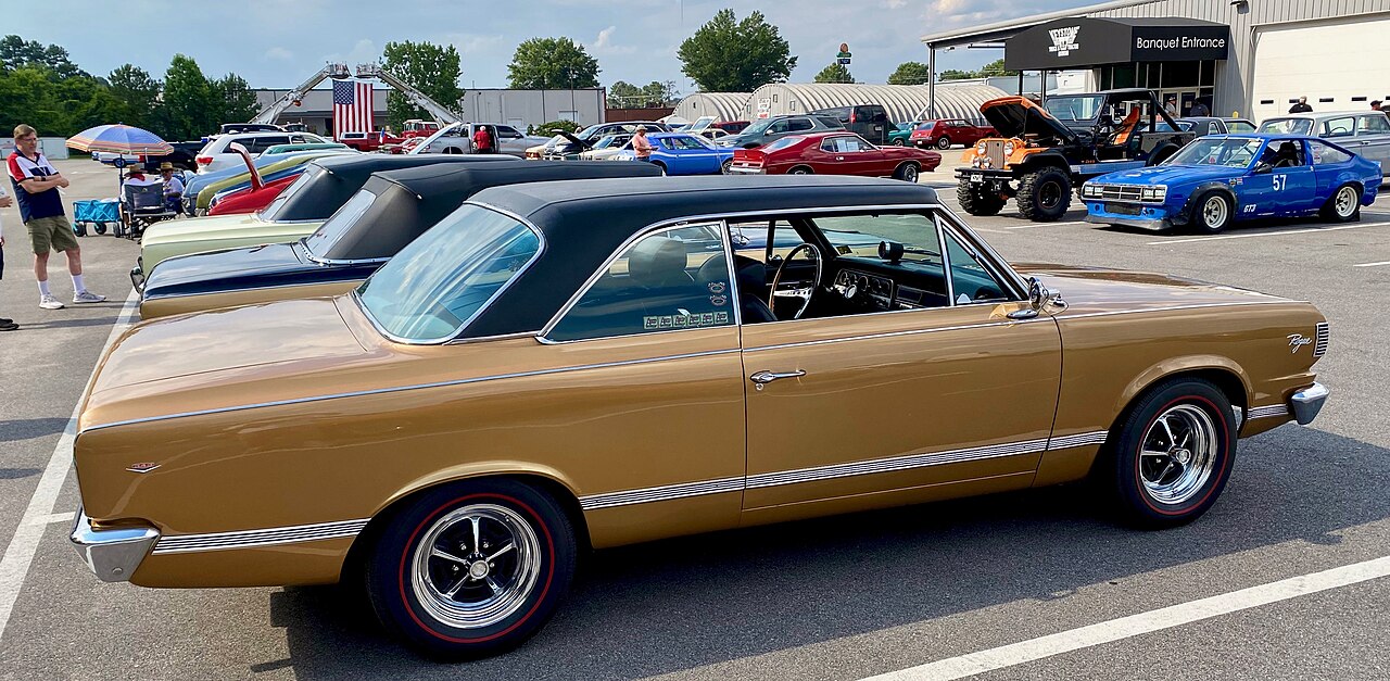 Gold classic muscle car with black vinyl roof and chrome wheels at outdoor car show with other vintage vehicles