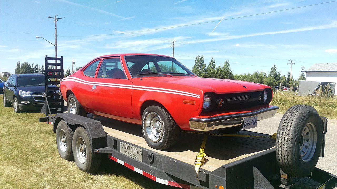 Red AMC Hornet hatchback with white racing stripes loaded on flatbed trailer with blue license plate in rural setting