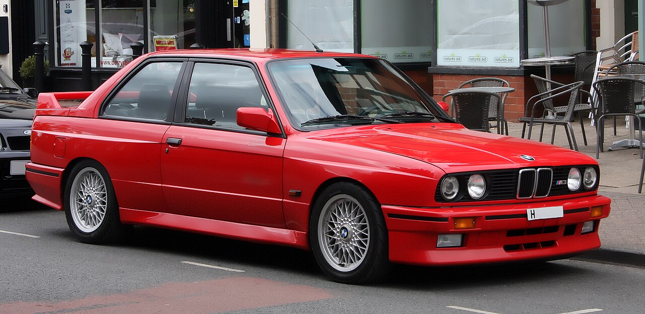 Red 1990 BMW M3 E30 with distinctive body kit and BBS wheels parked on urban street