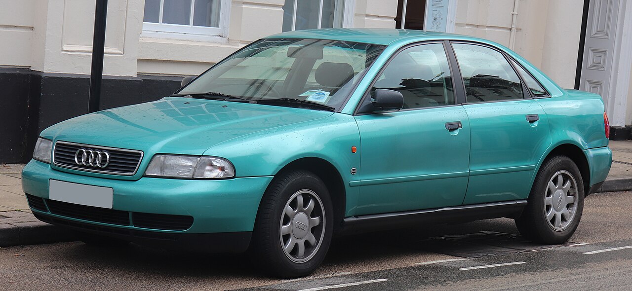 Teal green 1996 Audi A4 sedan with four-ring logo parked on street beside white building