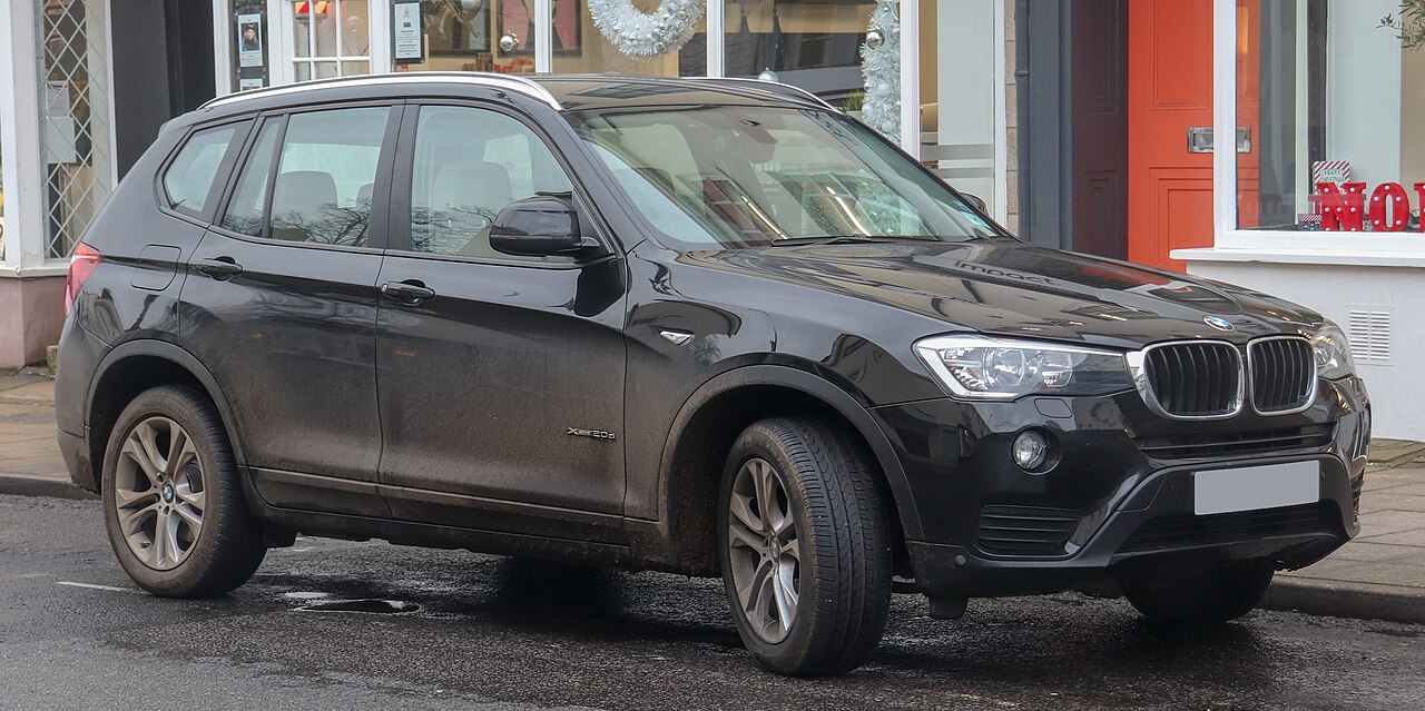 Dirty black BMW X3 SUV with mud and grime covering lower panels parked on street near storefronts