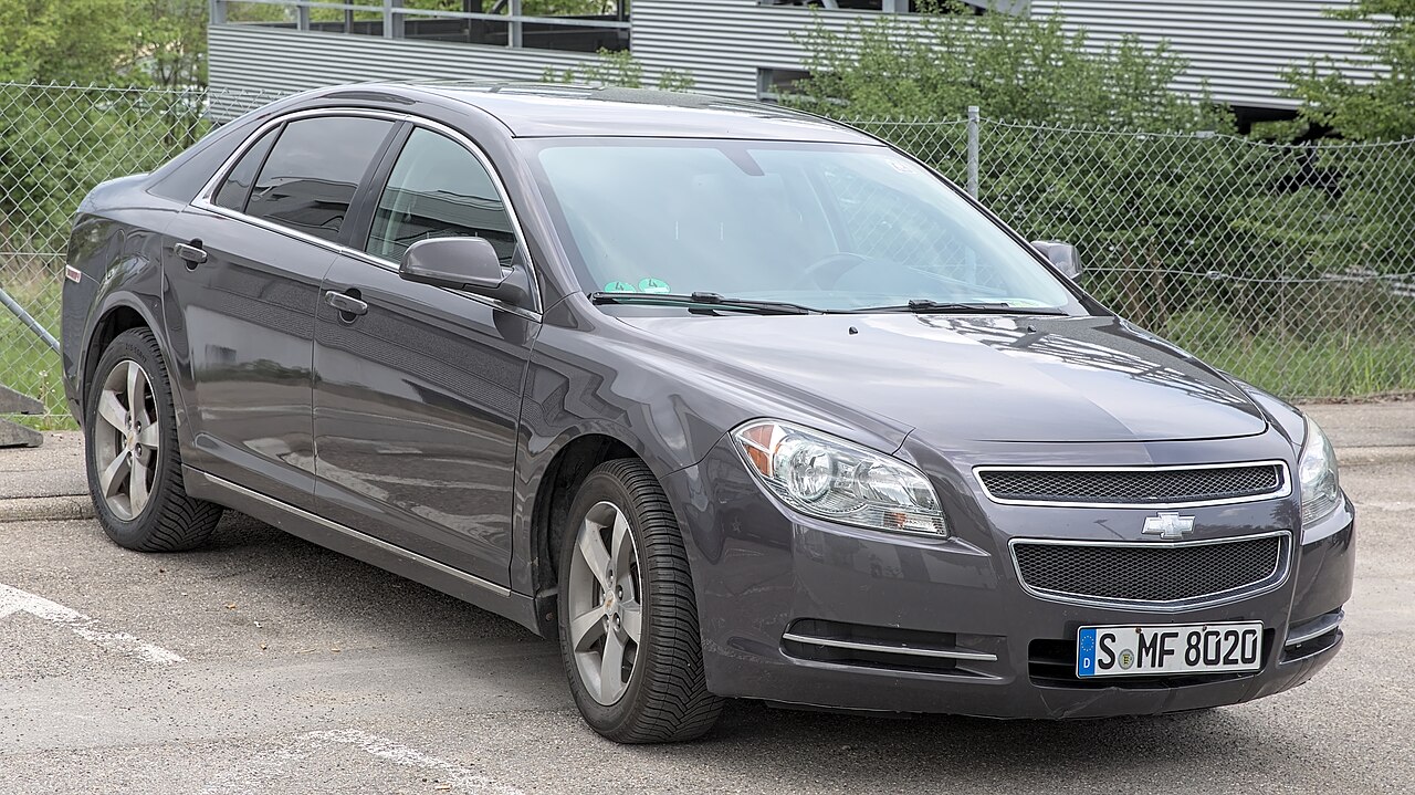 Black Chevrolet Malibu sedan parked on concrete with chain-link fence and industrial buildings in the background