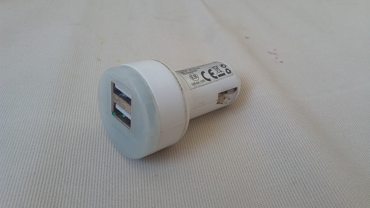 White dual-port USB car charger with CE certification markings lying on beige textured fabric surface