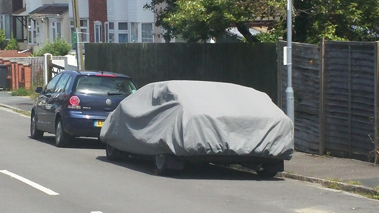Gray car cover protecting parked vehicle on residential street, with dark blue car nearby and houses in background