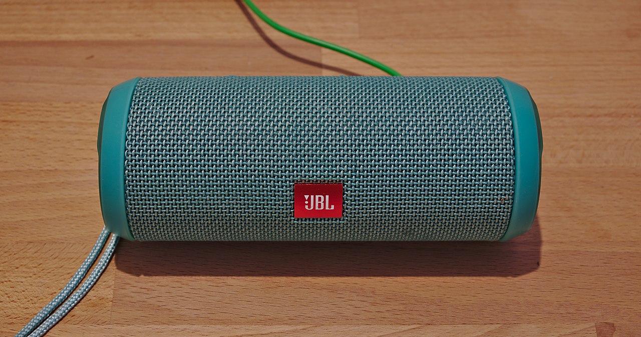 Teal JBL Bluetooth speaker with fabric mesh grille and red logo on wooden surface with charging cable