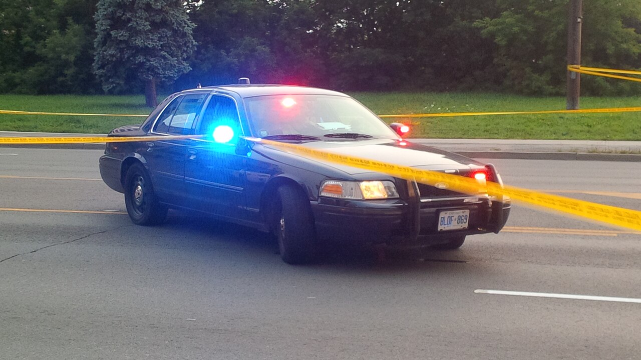 Dark unmarked police cruiser with activated blue and red emergency lights and spotlight, cordoned off with yellow tape