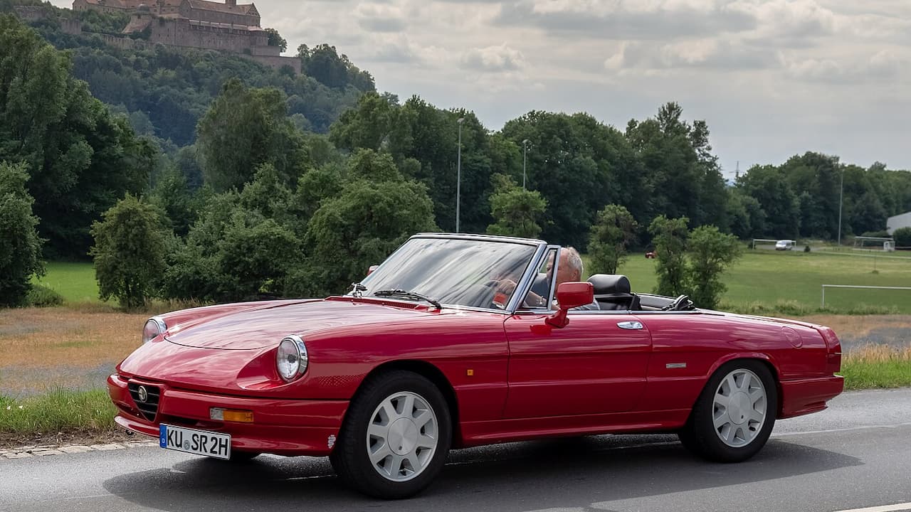 Red Alfa Romeo Spider with soft top parked on rural road with castle and countryside in background