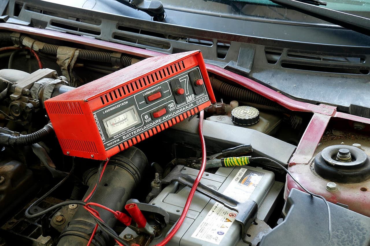 Red battery charger connected to car battery in engine compartment with cables, showing charging process for vehicle maintenance