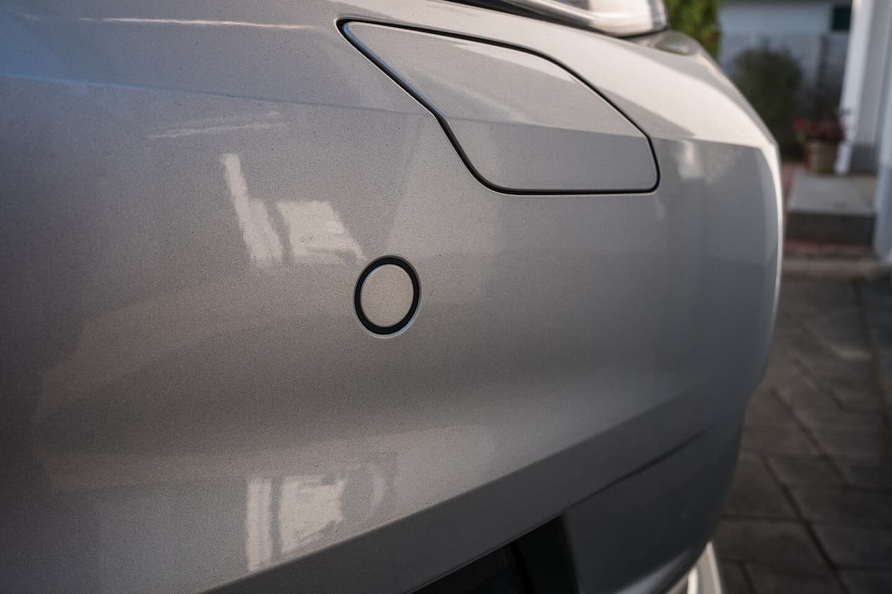Close-up of silver car's rear bumper showing circular parking sensor integrated into the vehicle's smooth metallic surface