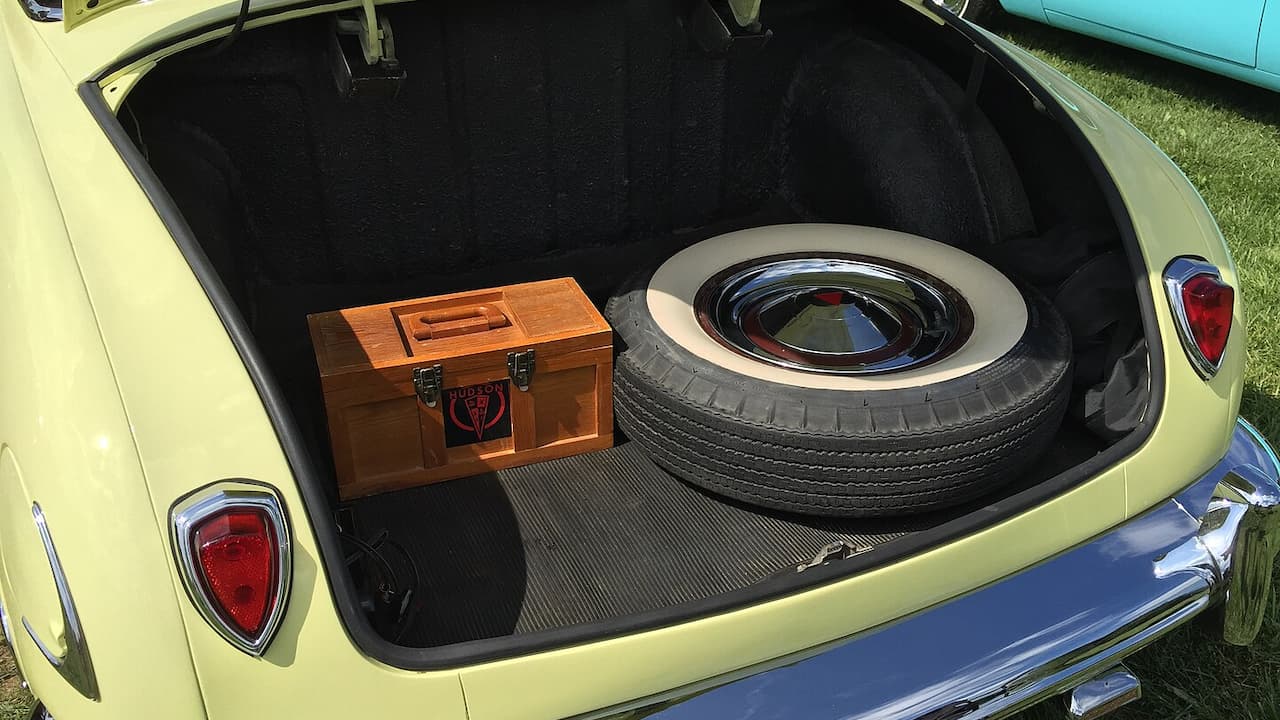 Open trunk of yellow classic car showing full-size spare tire with whitewall and wooden toolbox on black carpet